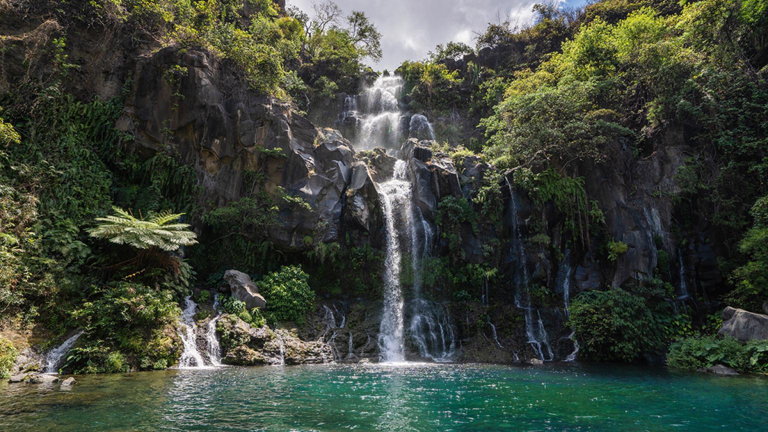 Full outdoor shot of a waterfall cascading down a rocky cliff face into a tranquil pool of emerald green water, Reunion
